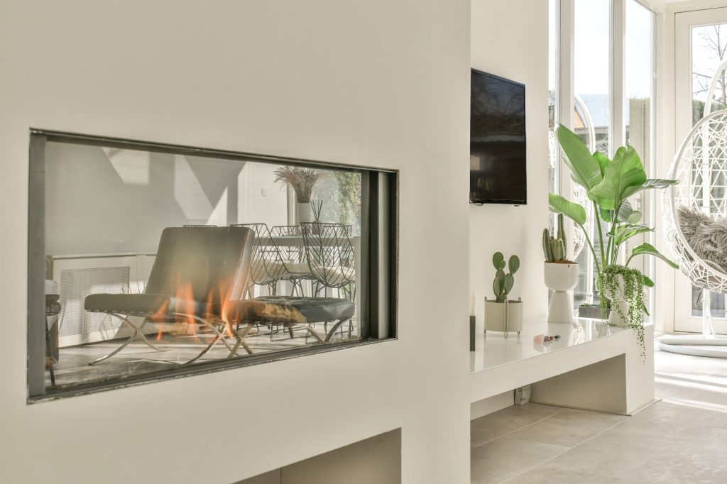 Bright sunroom with a lit see-through glass-front fireplace in a white wall, tropical plants and a hanging chair