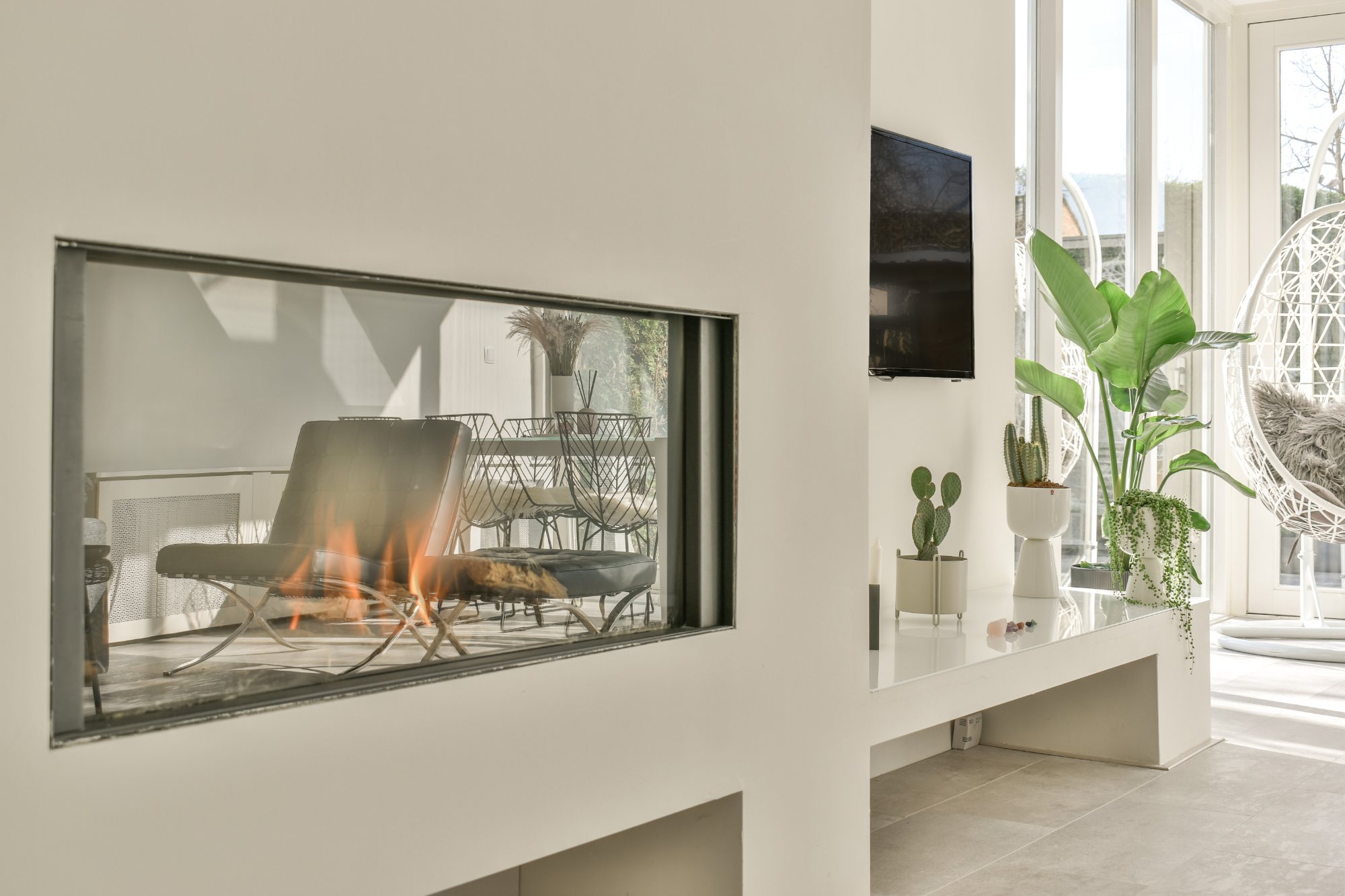 Bright sunroom with a lit see-through glass-front fireplace in a white wall, tropical plants and a hanging chair
