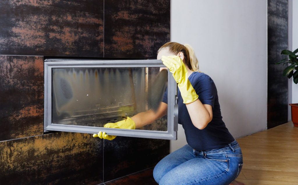Person in yellow rubber gloves wiping soot from the inside of a wood-burning fireplace's open glass door